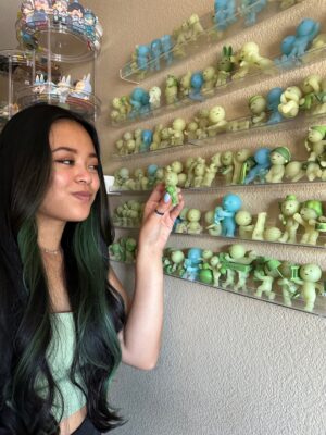 pacosalamander holds a green smiski and looks to her left at the rows of green and blue smiskis sitting on clear acrylic shelves on her wall. 