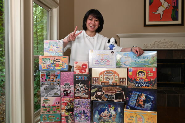 KiraKabuki makes a peace sign with her fingers and stands behind a chest-height stack of blind box cases with a smile.