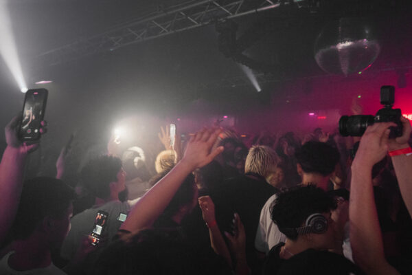 Dim room, with pink and white lighting, a crowd surrounds a blonde head, hair cut short.