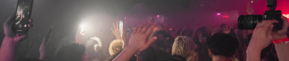 Dim room, with pink and white lighting, a crowd surrounds a blonde head, hair cut short.